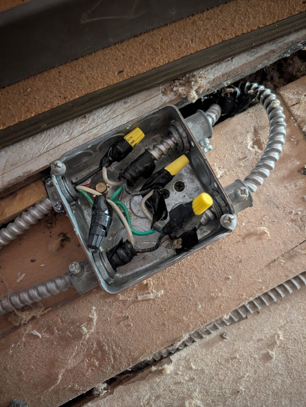 Open metal junction box between wooden joists, with flexible conduit and wire-nutted splices wrapped in black tape.